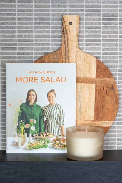 The cookbook 'More Salad' by Two Raw Sisters displayed on a dark countertop, styled with a wooden paddle board and a lit candle in front of a gray tiled wall.