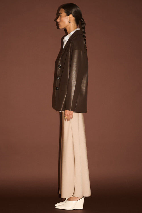 Person wearing a brown leather jacket and beige pants against a brown background