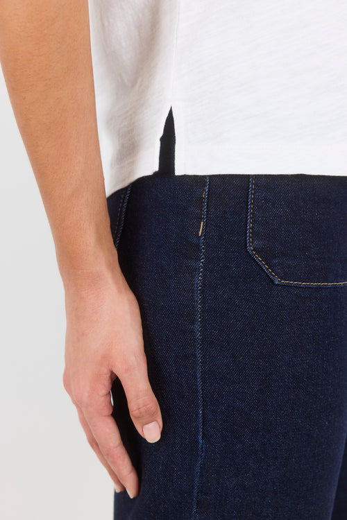 Close-up of a person wearing dark indigo denim jeans with a white top, showing the side pocket and hand area against a light gray background.