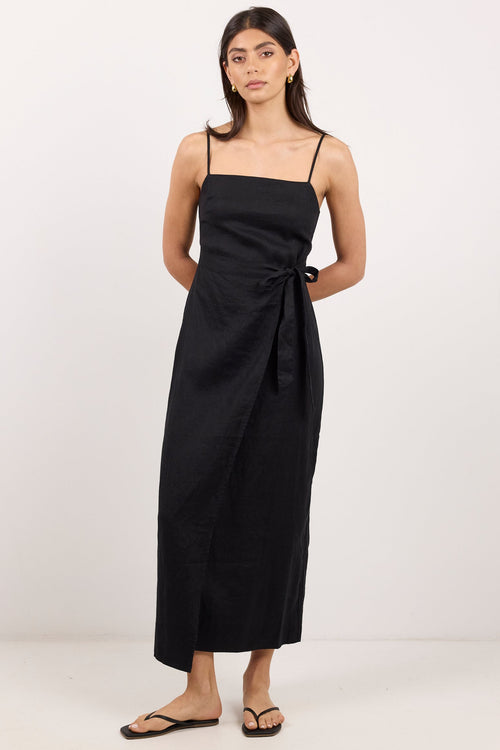 A woman modeling a black midi wrap dress with spaghetti straps and a side tie detail, paired with black flip-flop sandals, against a plain white studio background.