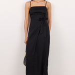 A woman modeling a black midi dress with thin straps, a square neckline, and a side tie detail, paired with black flip-flop sandals against a plain white studio background.