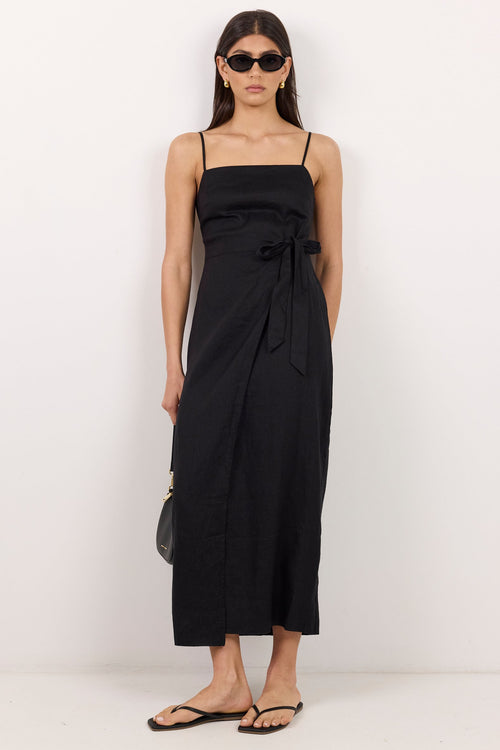 A woman modeling a black midi dress with thin straps, a square neckline, and a side tie detail, paired with black flip-flop sandals against a plain white studio background.