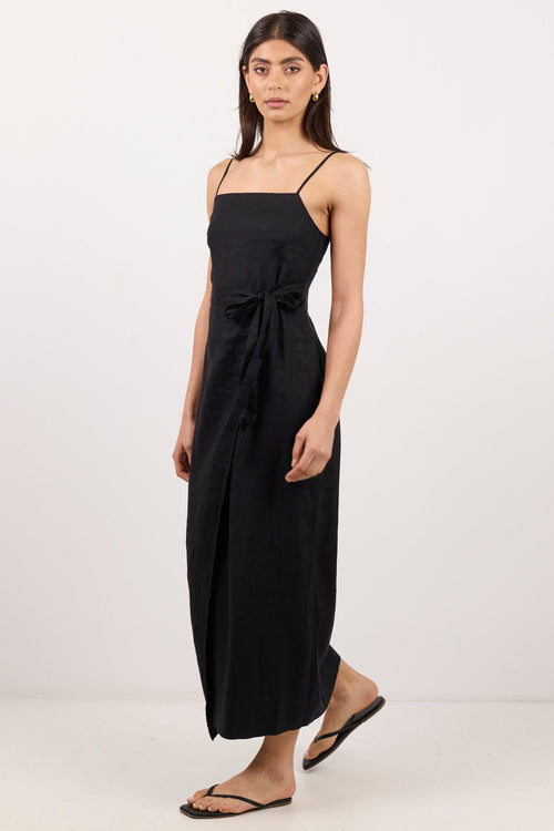 A woman modeling a black midi-length spaghetti-strap dress with a tied waist detail, paired with black flip-flop sandals, against a plain white studio background.