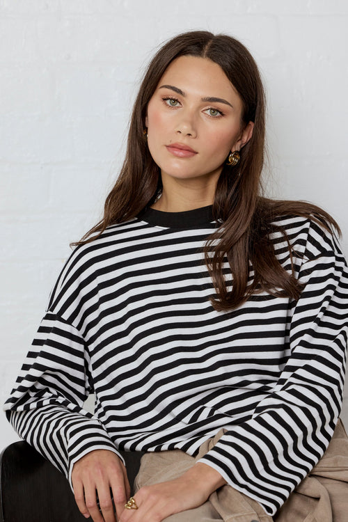 A woman wearing a black and white horizontally striped long-sleeve top, paired with beige pants, sitting against a white brick wall.