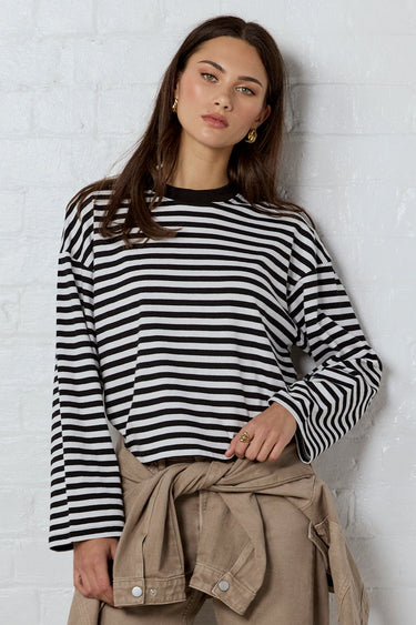 A woman stands against a white brick wall wearing a black and white striped long-sleeve top, beige cropped pants with a matching tie belt, and black flat shoes.