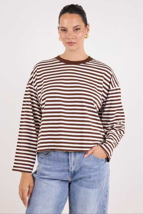 A woman modeling a brown and white horizontally striped long-sleeve crewneck top with light blue jeans, standing against a plain light gray studio background.