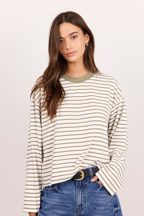 A woman modeling a long-sleeve crewneck top with thin beige and white horizontal stripes, paired with blue jeans and a black belt, against a plain light background.