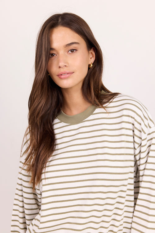 A woman with long brown hair wearing a cream and olive green horizontally striped long-sleeve shirt, photographed against a plain light background.