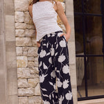 Woman wearing a white sleeveless top and navy floral pants leaning against a stone wall.