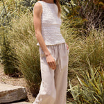 Woman in a white sleeveless top and light pants standing in a garden with greenery and purple flowers.