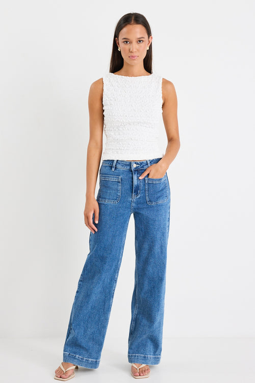 Woman wearing a white sleeveless textured top with a fitted silhouette and high neckline, paired with high-waisted denim jeans.