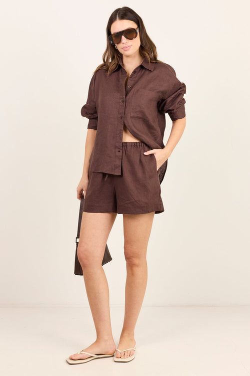 A woman modeling a matching dark brown linen shirt and shorts set, styled with sunglasses, a black shoulder bag, and beige flip-flops against a plain off-white studio background.