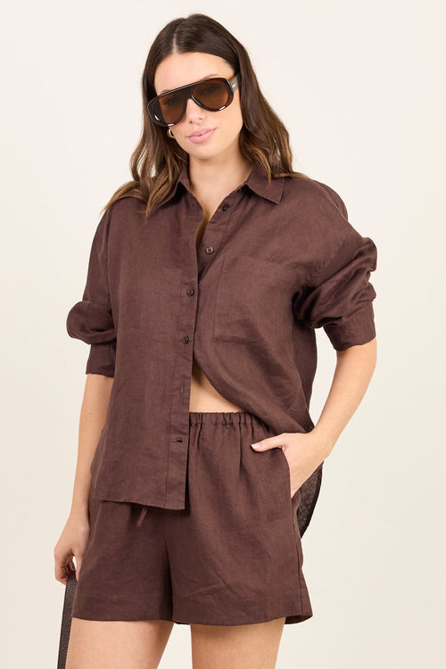 A woman modeling a matching dark brown linen shirt and shorts set, styled with sunglasses, a black shoulder bag, and beige flip-flops against a plain off-white studio background.