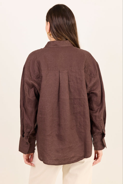 A woman viewed from the back wearing a brown long-sleeve linen shirt and cream pants, standing against a plain off-white background.