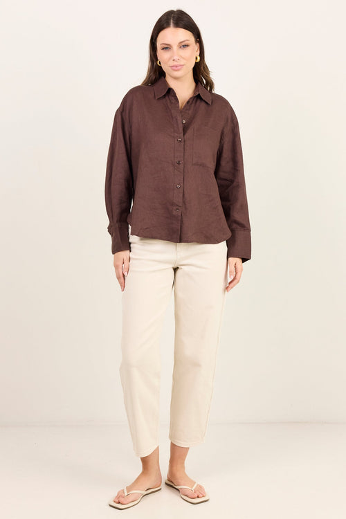 A woman modeling a dark brown long-sleeve button-up shirt and cream cropped trousers, standing against a plain off-white studio background.