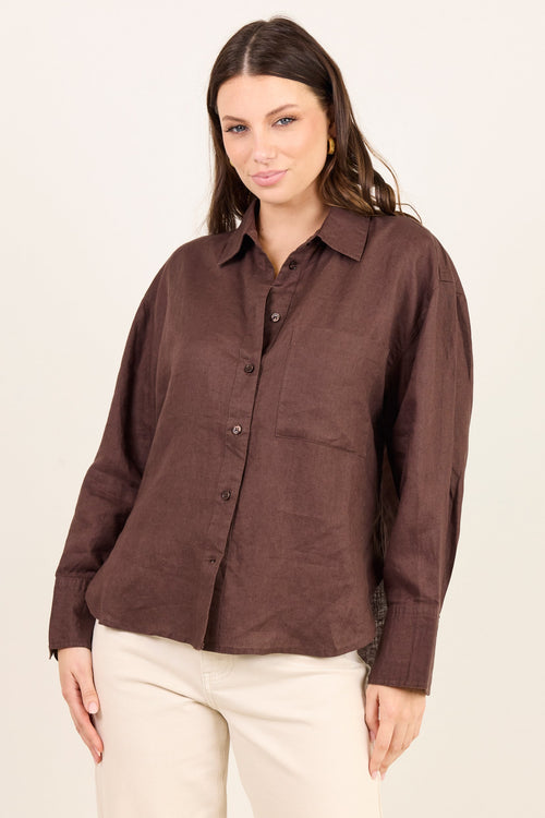 A woman modeling a brown long-sleeve linen button-up shirt with cream pants, photographed against a solid off-white studio background.