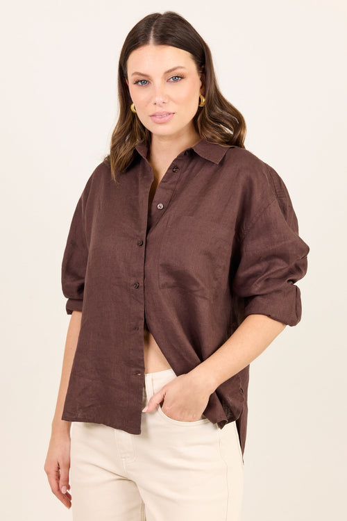 A woman modeling a dark brown linen button-up shirt with rolled-up sleeves, paired with cream pants, against a plain off-white background.