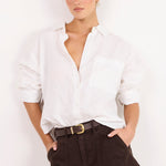 A woman modeling a white long-sleeve button-down shirt and dark brown shorts with a black belt, standing against a plain white background.