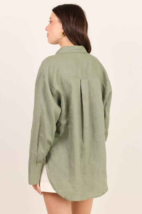 A woman shown from the back wearing an oversized sage green linen button-up shirt, paired with a light beige skirt, against a solid off-white background.