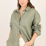 A woman modeling an oversized sage green linen button-up shirt with cream shorts, photographed against a solid off-white studio background.