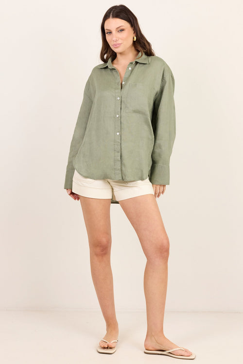 A woman modeling a sage green long-sleeve linen button-up shirt with off-white shorts and beige sandals, standing against a plain off-white studio background.