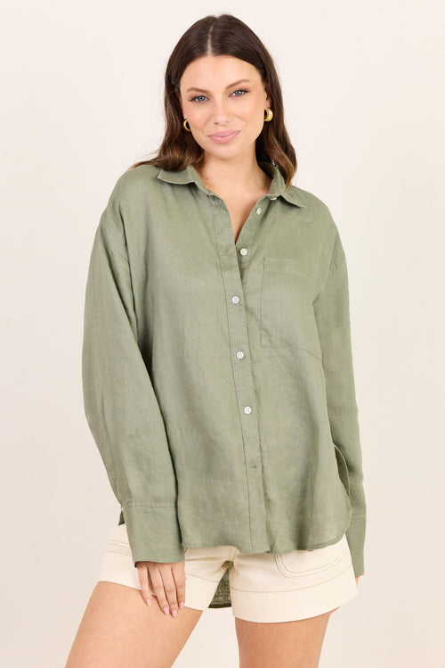 A woman modeling an oversized sage green linen button-up shirt with cream shorts, set against a plain off-white studio background.