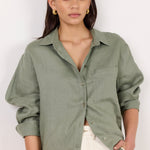 A woman modeling an oversized sage green linen button-up shirt with rolled sleeves and white shorts, set against a plain white studio background.