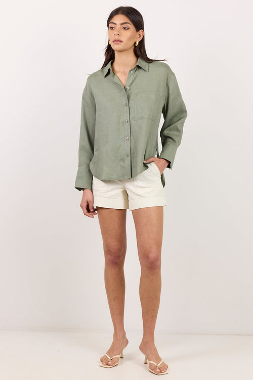 A woman modeling a sage green long-sleeve linen button-up shirt paired with off-white shorts and beige sandals, standing against a plain white studio background.