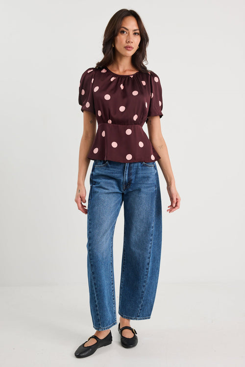 Woman wearing a plum-coloured short sleeve top with blush pink polka dots, gathered neckline, and a cinched waist, styled with blue jeans.