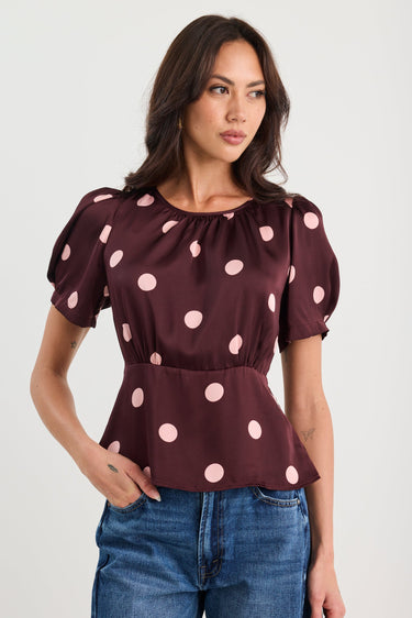 Woman wearing a plum-coloured short sleeve top with blush pink polka dots, gathered neckline, and a cinched waist, styled with blue jeans.