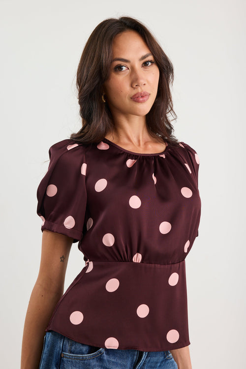 Woman wearing a plum-coloured short sleeve top with blush pink polka dots, gathered neckline, and a cinched waist, styled with blue jeans.