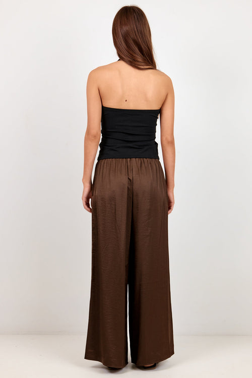 A woman viewed from behind wearing a black strapless top and brown wide-leg pants against a plain light gray studio background.