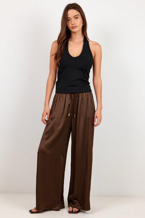 A woman modeling wide-leg brown pants with a drawstring waist, paired with a black tank top, standing against a plain white studio background.