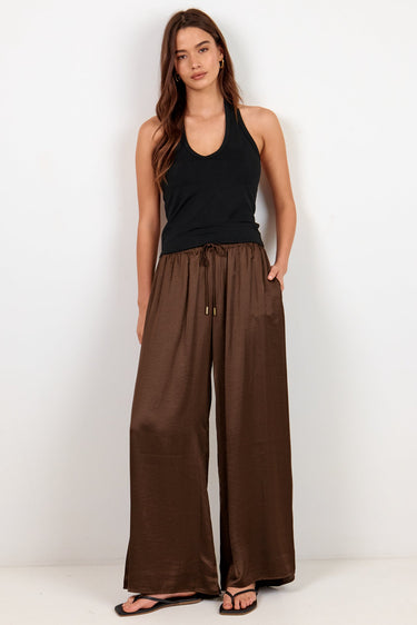A woman modeling wide-leg brown pants with a drawstring waist, paired with a black tank top and black sandals, standing against a plain white studio background.