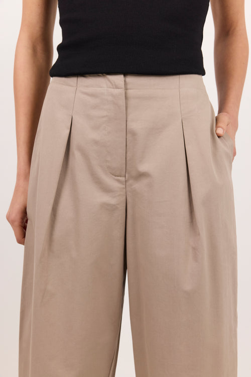 A close-up of a model wearing high-waisted, wide-leg pleated trousers in a light taupe-beige color, paired with a black top against a plain light background.