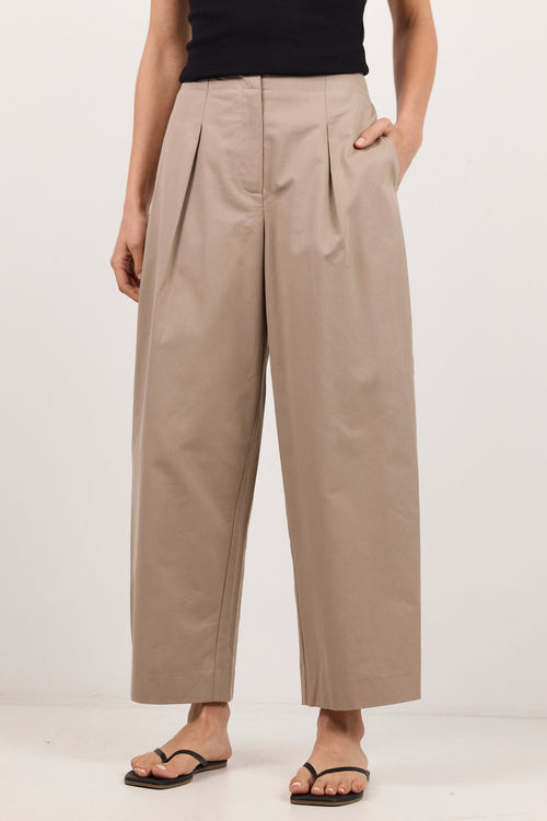 A model wearing high-waisted, wide-leg cropped trousers in a light taupe-beige color, paired with a black sleeveless top and black flip-flop sandals, against a plain white studio background.