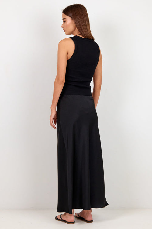 A woman stands with her back to the camera wearing a sleeveless black top and a long black skirt against a plain white studio background.