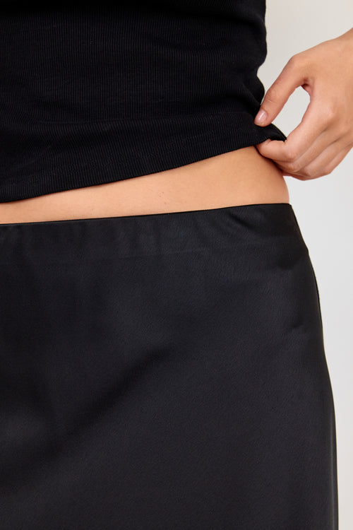 Close-up of a person wearing a black ribbed top and black skirt, lifting the hem of the top to show the waistband, against a light gray background.
