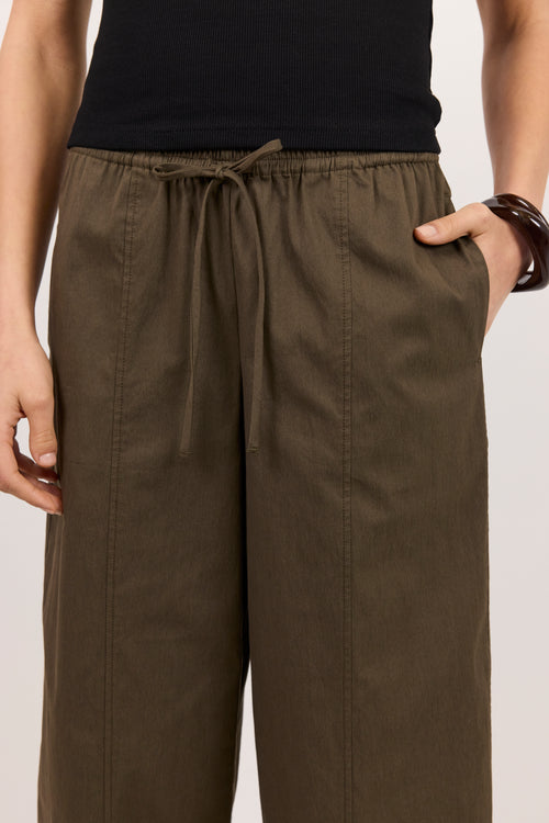 A close-up of a person wearing wide-leg olive brown drawstring pants with a black top, standing against a light neutral background.