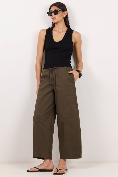 A woman modeling wide-leg cropped pants in olive green, paired with a black tank top, black sunglasses, and black flip-flops, standing against a plain white studio background.