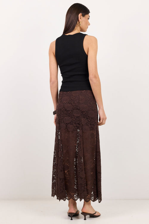 A woman shown from the back wearing a sleeveless black top and a long brown lace skirt, standing against a plain white studio background.