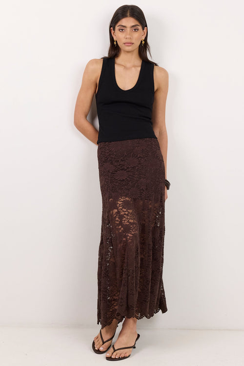 A woman stands against a plain white studio wall wearing a black sleeveless top and a long dark brown lace maxi skirt with a draped side detail.