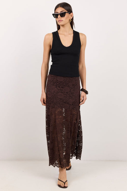 A woman modeling a sleeveless black ribbed tank top and a dark brown lace maxi skirt, styled with black sunglasses and flat sandals, against a plain white studio background.
