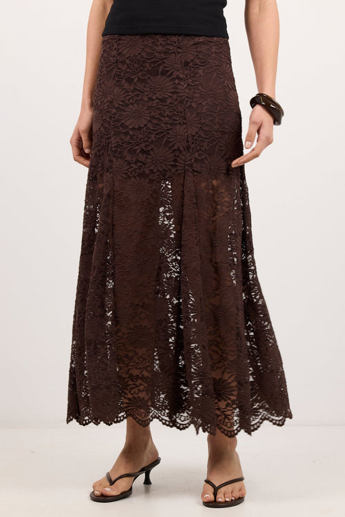 A model wearing a dark brown lace maxi skirt with a scalloped hem, paired with a black top and black sandals, against a plain white studio background.