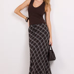 A woman modeling a dark brown ribbed tank top and a long black and white windowpane plaid maxi skirt, holding a black handbag against a plain white studio background.
