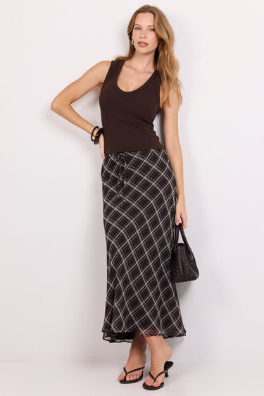 A woman modeling a dark brown ribbed tank top and a long black and white windowpane plaid maxi skirt, holding a black handbag against a plain white studio background.