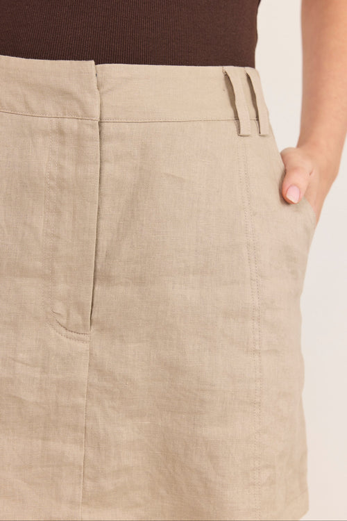 Close-up of a beige linen skirt with a side pocket and belt loops, worn with a dark brown ribbed top against a light neutral background.