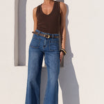 A woman stands outdoors against a white wall wearing a brown sleeveless top, blue wide-leg jeans, a brown belt, and black sandals.