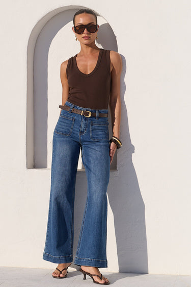 A woman stands outdoors against a white wall wearing a brown sleeveless top, blue wide-leg jeans, a brown belt, and black sandals.