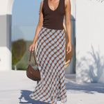 A woman walking outdoors near a white architectural structure and pool, wearing a sleeveless dark brown top and a long brown-and-beige checkered skirt, with sunglasses and a brown handbag.
